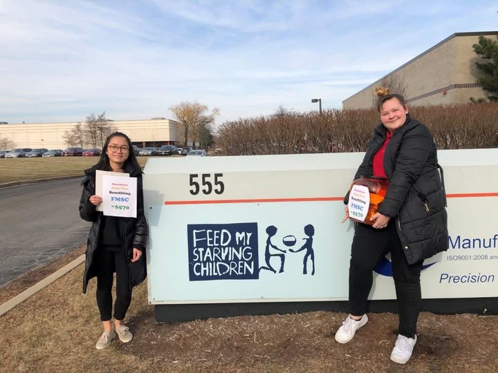 Two young ladies stand infront of feed my starving children, holding posters that signify the money they raised for this organization. The background shows a blue sky and it is mid-fall. 