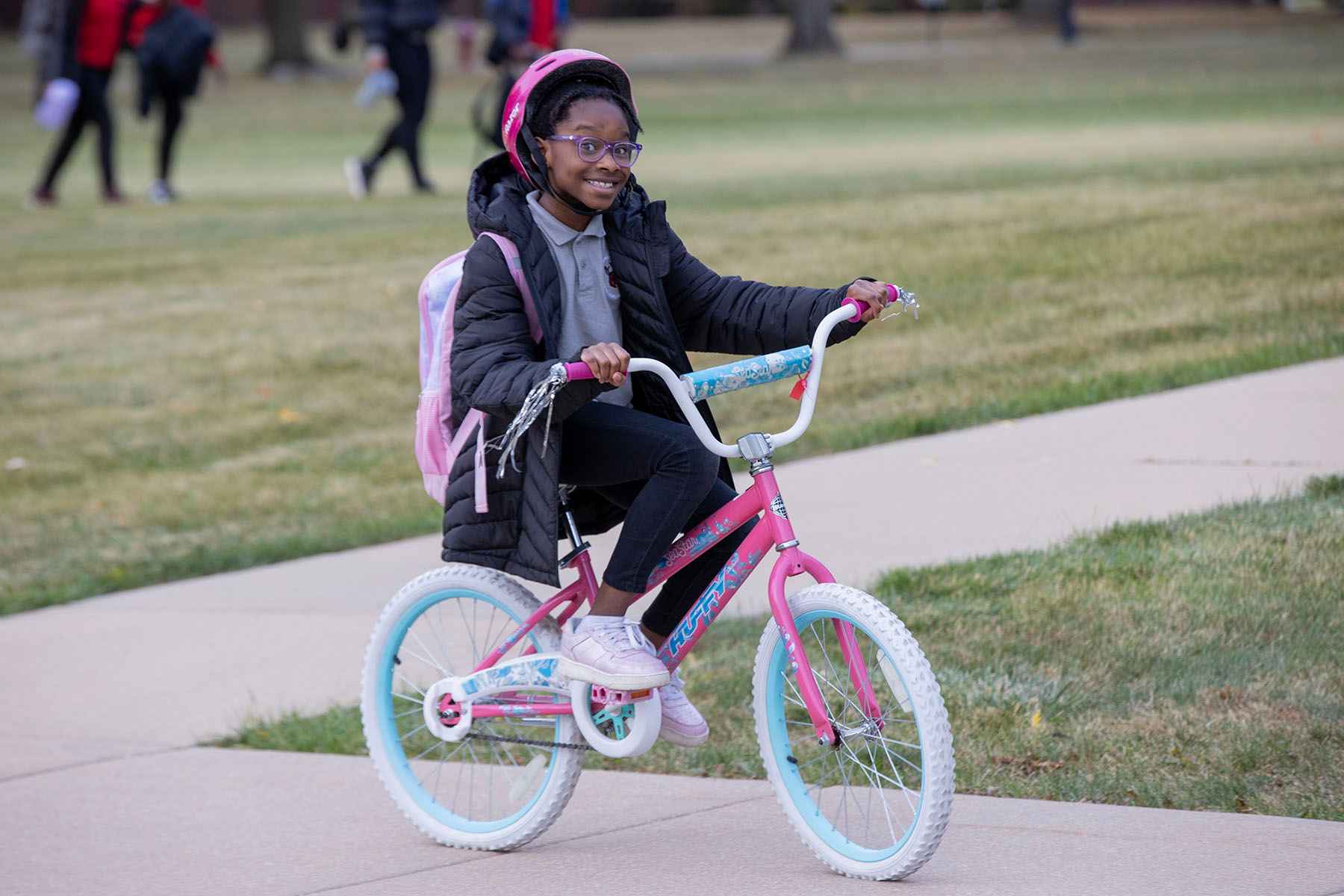 A young girl pedaling her bike along a sidewalk, with trees and a sunny sky in the background.
